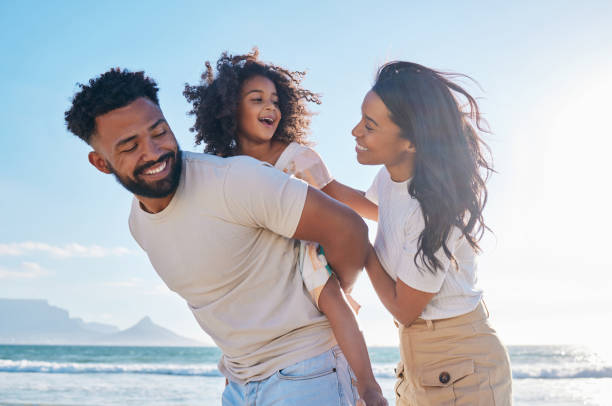 Família de três pessoas. O pai carrega a filha nas costas e todos sorriem felizes e tranquilos em uma praia